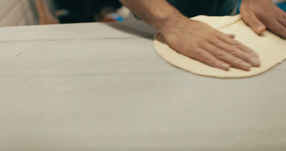 Two hands pressing and stretching a round of raw pizza dough flat on a floured bench.