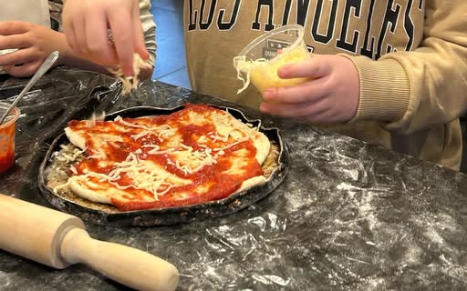 Children's hands scattering mozzarella over a sauced pizza base on a black pan at a Numero Uno pizza-making party.