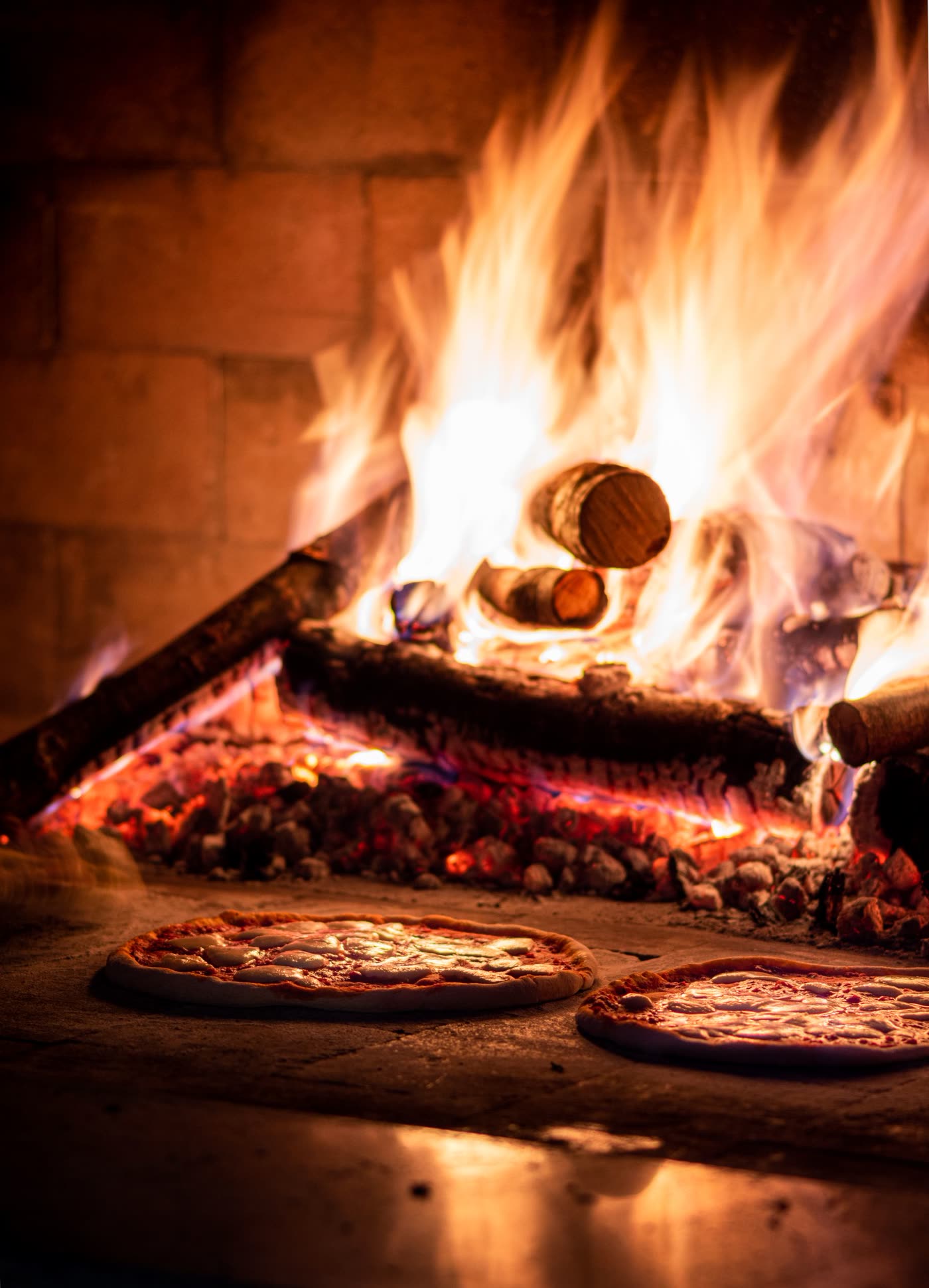 Two pizzas cooking inside a stone wood-fired oven beside an open flame.