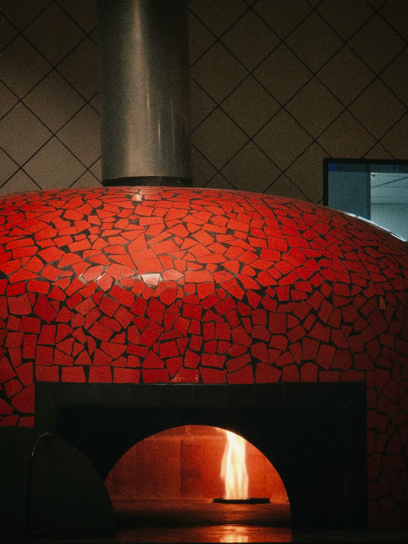 A rustic wood-fired pizza oven faced in red tile mosaic with a glowing interior.
